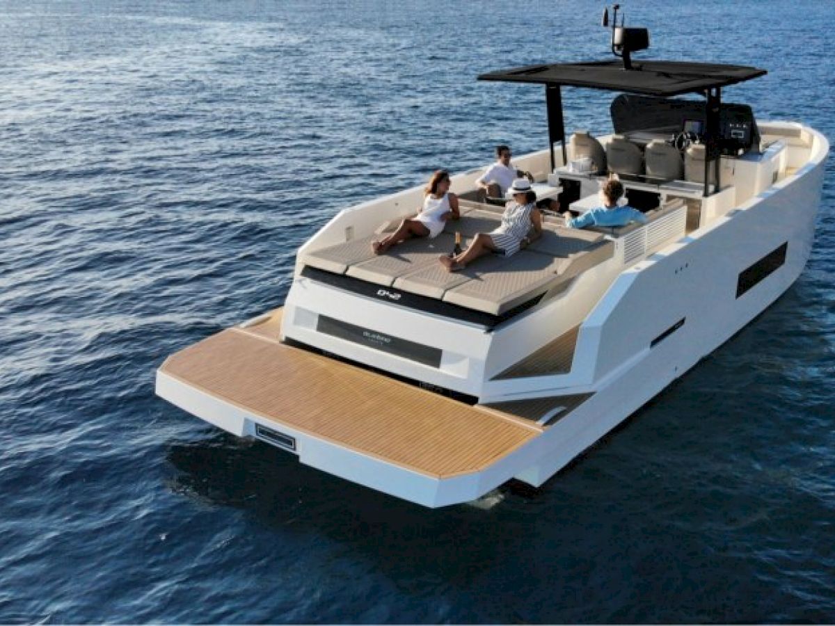 A modern yacht is on the water, with four people lounging at the back. The sea is calm and the sky is clear.