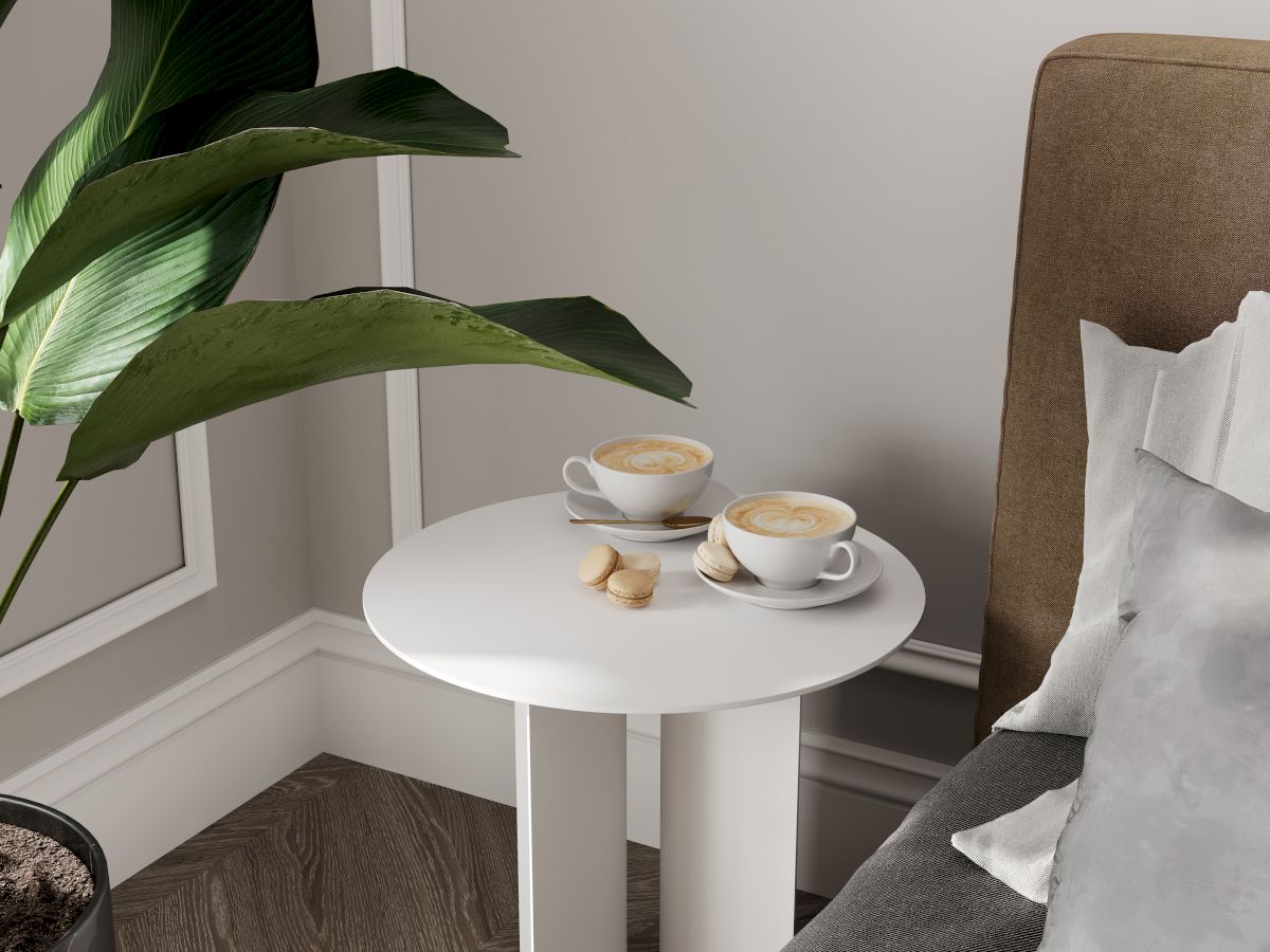 A small white table beside a bed holds two cups of coffee and cookies, with a large green plant nearby.