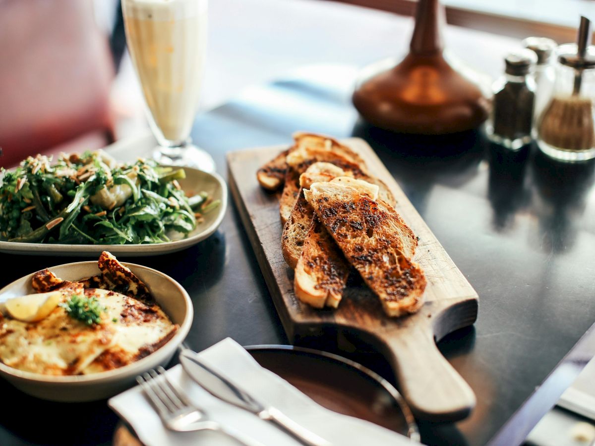 The image shows a meal with toasted bread on a wooden board, a green salad, a baked dish, and a drink on a table.