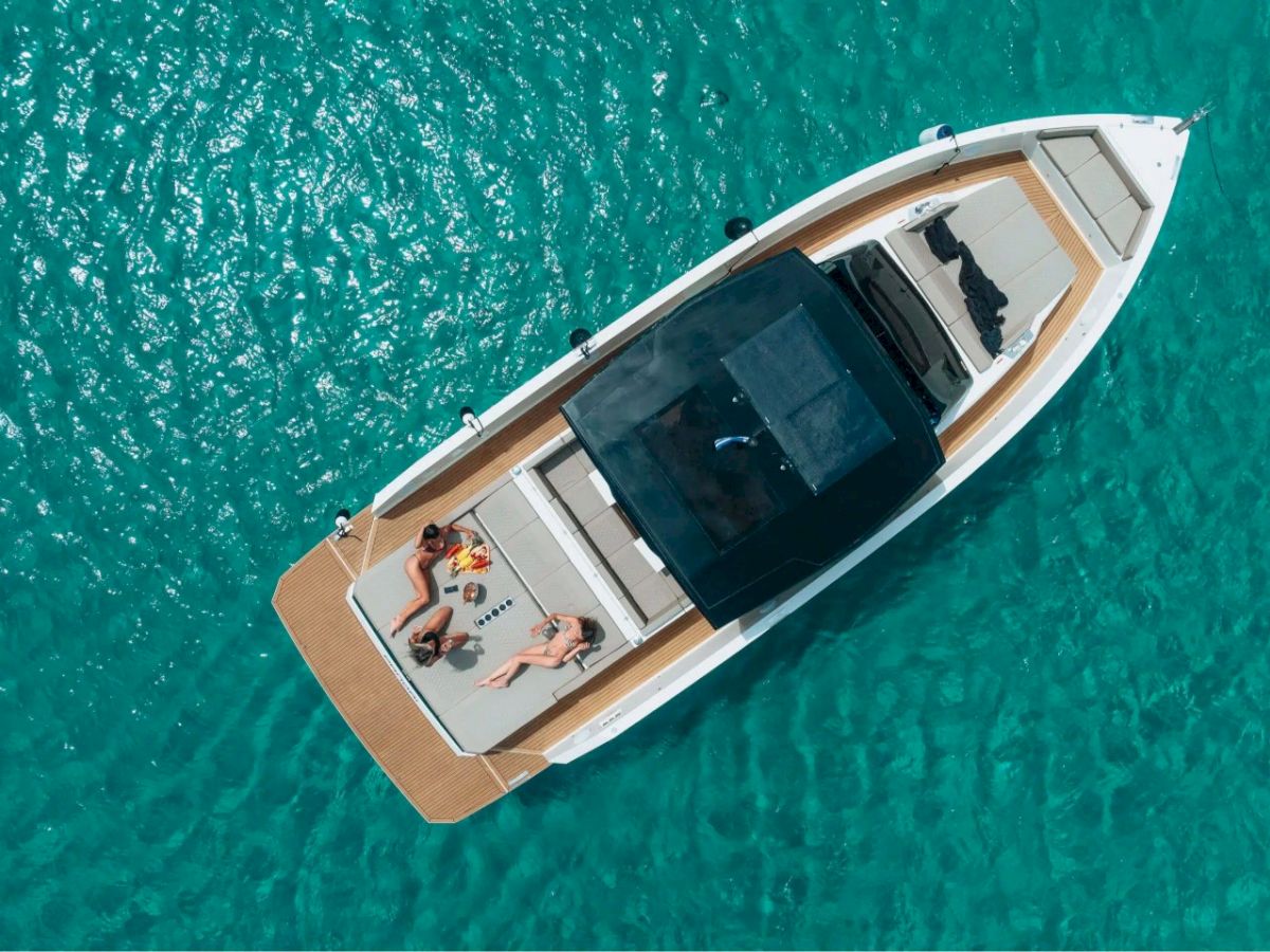 Aerial view of a boat on clear blue water with two people relaxing on the deck under the sun, enjoying a leisure day on the sea.