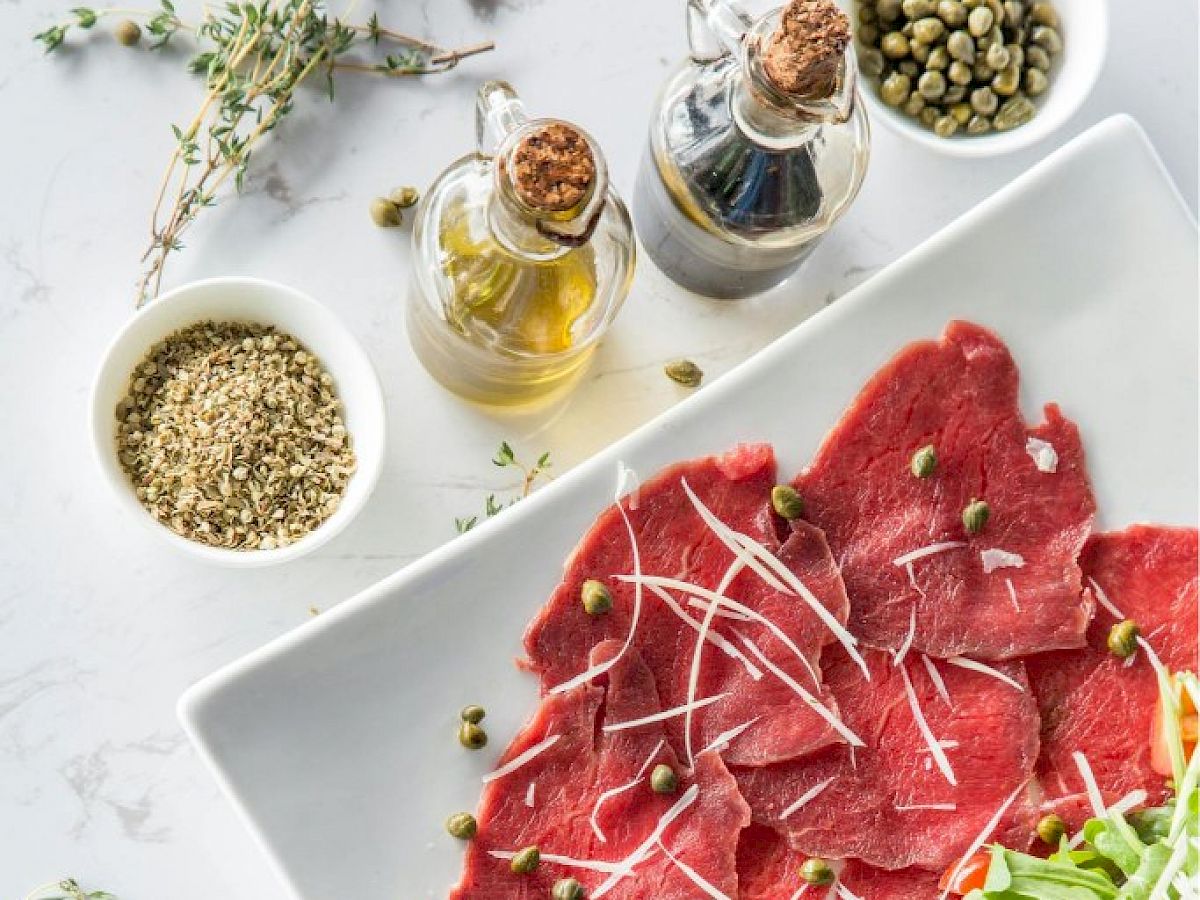 The image shows a plate of beef carpaccio with arugula and cheese shavings, plus capers, herbs, olive oil, and vinegar on the side.