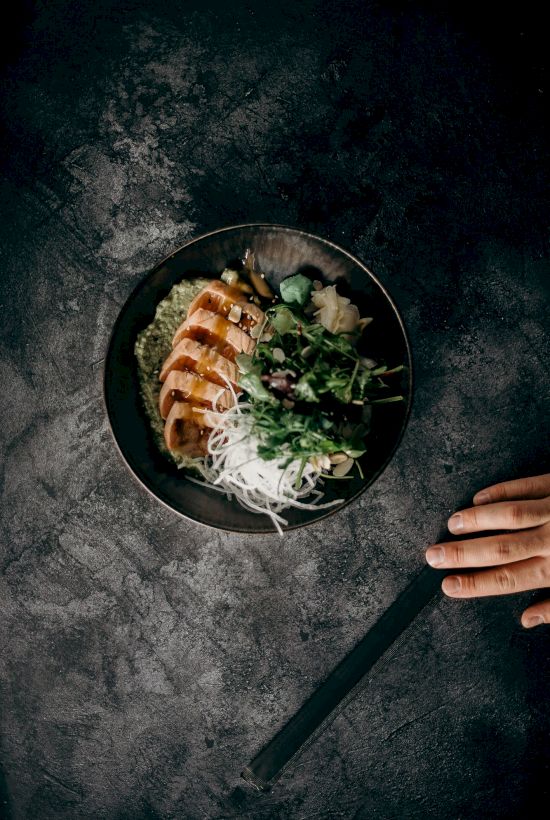 A bowl of grilled meat, noodles, and greens sits on a dark surface, with a hand and chopsticks nearby.