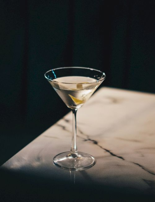 A martini glass with a clear cocktail and a lemon twist sits on a marble surface, illuminated under a low light.