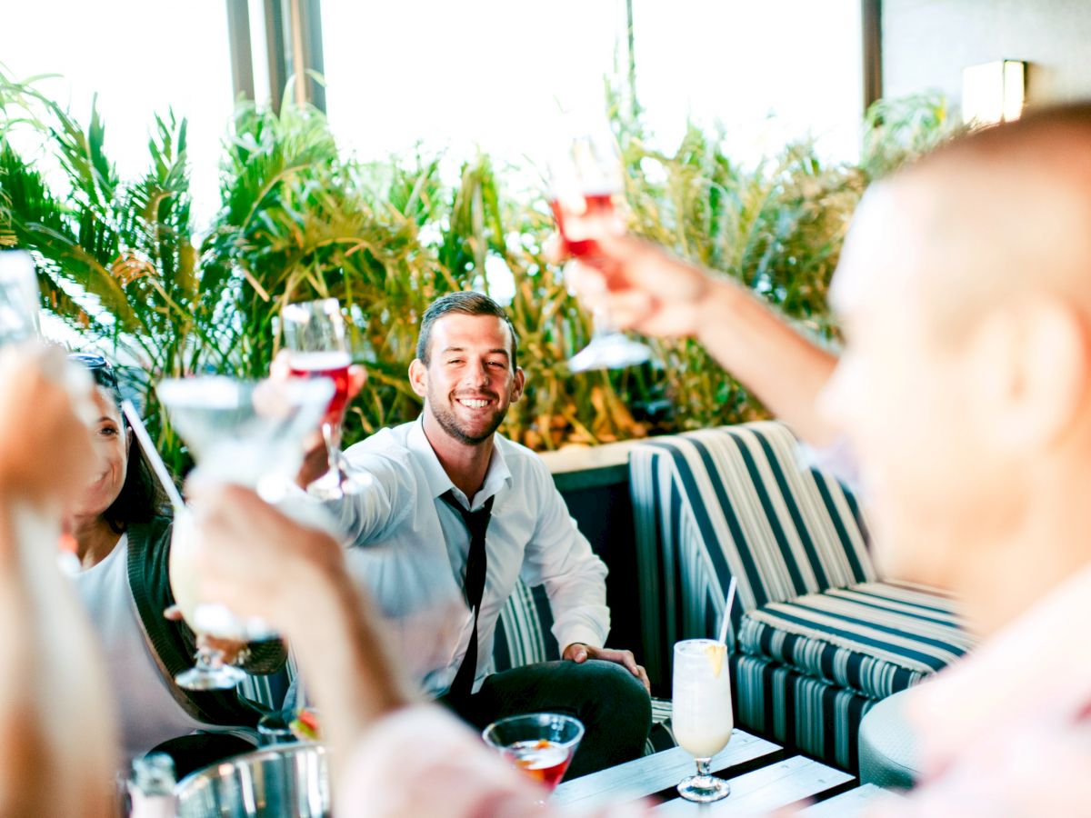 People are raising glasses in a toast, seated in a casual setting with greenery in the background, creating a lively atmosphere.